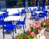 Terasa kavárny s modrými židlemi a bílými stoly vedle květináče s růžovými květinami.