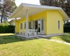 Žlutý bungalov s terasou a plastovými židlemi, obklopený trávníkem a stromy.  