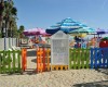 Dětské hřiště na pláži obklopené barevným plotem a slunečníky.