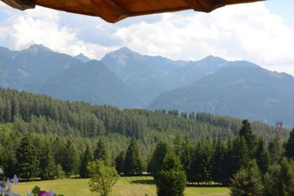 Zalesněná krajina s horským pohořím v pozadí a dřevěným okrajem střechy nahoře.
