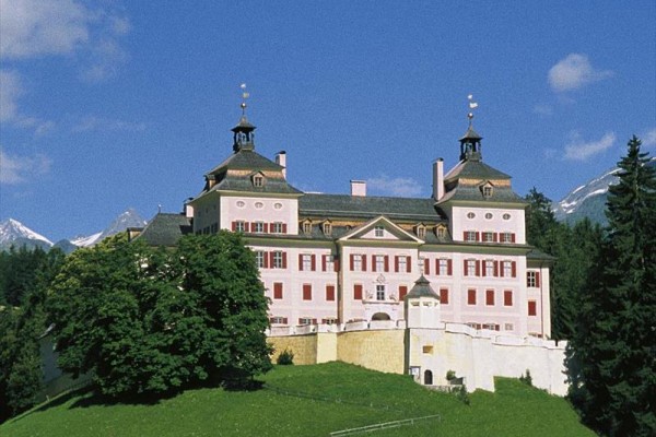 Historický zámek s věžičkami obklopený zelení a horami.