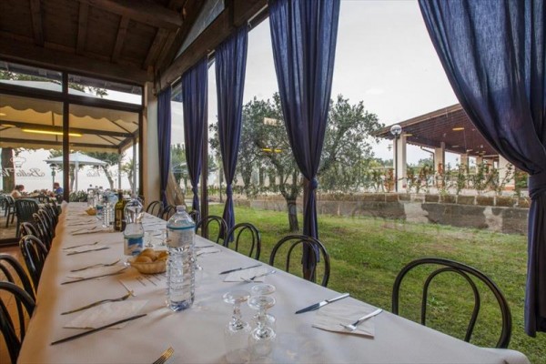 Dlouhý prostřený stůl v restauraci poblíž velkých oken se závěsy, s výhledem na venkovní terasu a zahradu.
