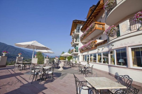 Venkovní terasa u hotelu s kovovými stolky a židlemi, obklopená květinami na balkonech.