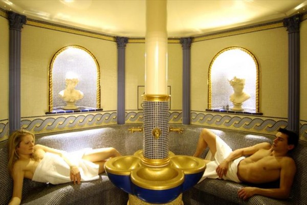 Dva lidé relaxují na lehátkách v luxusní místnosti s mozaikovou výzdobou.