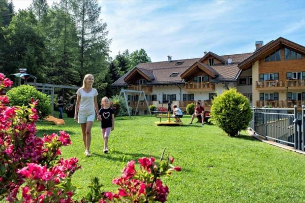 Žena s dítětem procházejí zahradou poblíž dřevěných budov, v pozadí hřiště.