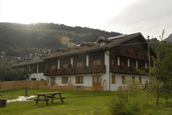 Penzion s dřevěnými balkony a lavičkou v popředí na pozadí kopců.