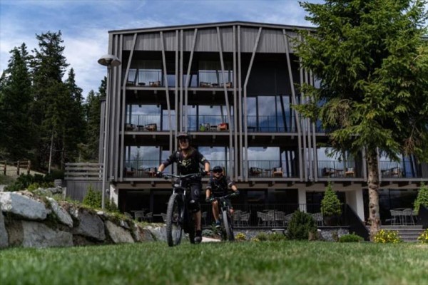 Dva cyklisté jedoucí na horských kolech před moderní budovou obklopenou stromy.