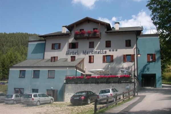 Třípatrový hotel s balkony ozdobenými květinami a několika parkovanými auty před budovou.