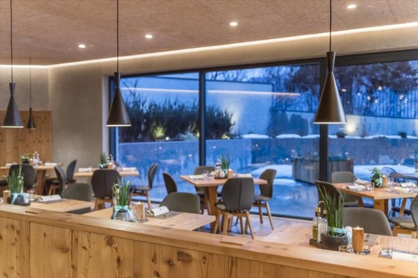 Moderní restaurace s dřevěnými stoly, židlemi a velkými okny s výhledem ven.