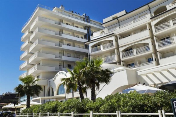 Moderní hotel s balkony a palmami v popředí za modré oblohy.