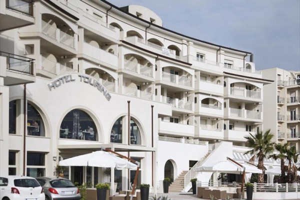 Moderní hotel se světlou fasádou a venkovní terasou s bílými slunečníky.