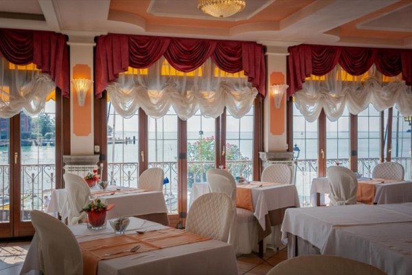 Elegantní restaurace s bílými ubrusy, květinovou výzdobou a výhledem na jezero přes velká okna.