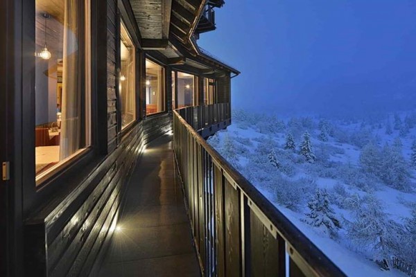 Úzký dřevěný balkon vedle osvětlené restaurace s výhledem na zasněženou krajinu večer.