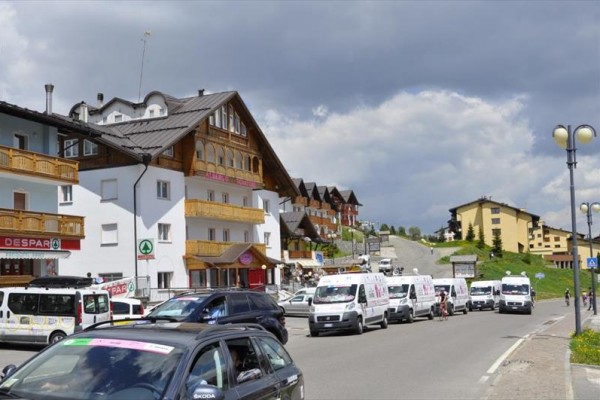 Ulice s několika automobily a domy s dřevěnými balkony pod zataženou oblohou.