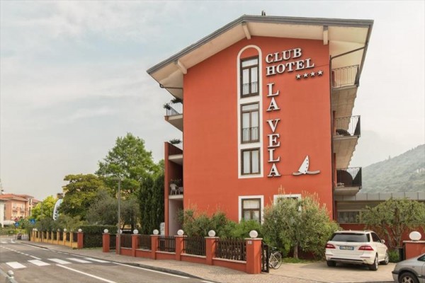 Červený budova hotelu Club Hotel La Vela se čtyřmi hvězdičkami a parkovištěm před ní.