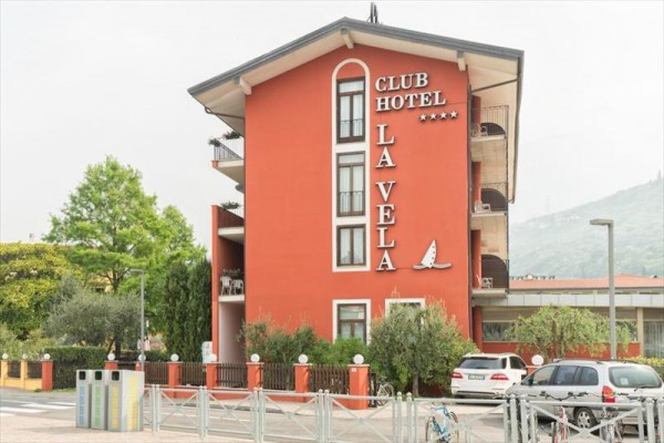 Červená budova hotelu La Vela s balkonem, obklopená zelení a zaparkovanými auty.
