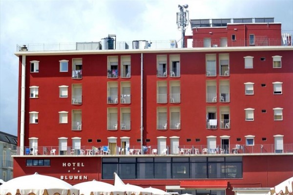 Cihlově červená fasáda hotelu s balkóny a venkovním posezením.