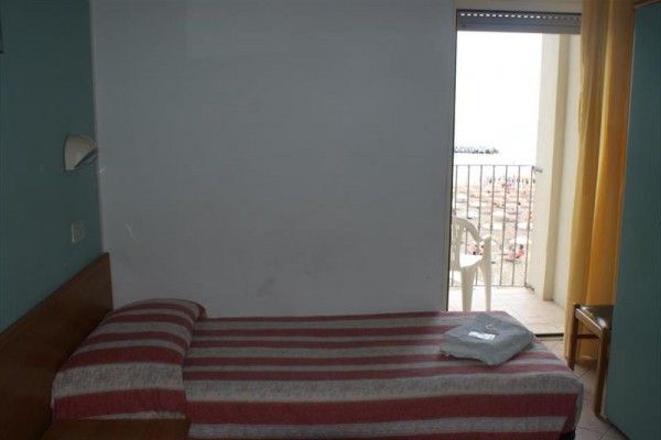 Pokoj s úzkou postelí a výhledem na balkón směrem k pláži.