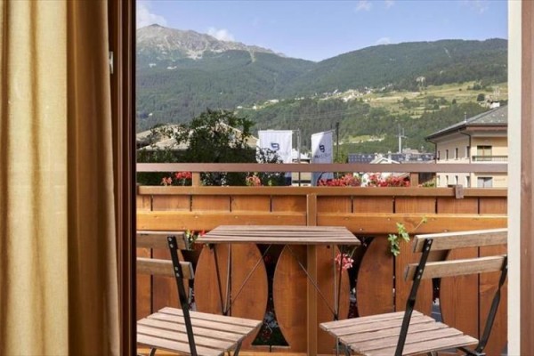 Výhled z pokoje na balkon se stolem a židlemi, v pozadí hory.