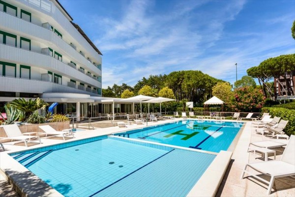 Venku je bazén s lehátky vedle moderního hotelu obklopeného zelení.  