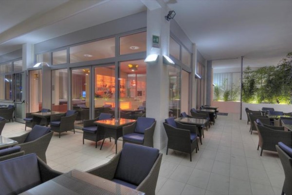 Elegantní venkovní terasa restaurace s pohodlnými křesly a stolky, za skleněnými dveřmi osvětlený interiér.  