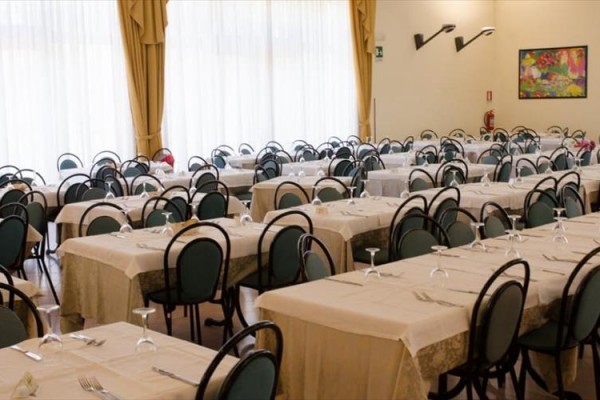 Restaurace s řadami prostřených stolů a židlemi před velkým oknem se závěsy.
