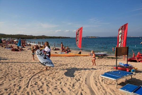Slunečná pláž plná lidí, s červenými plachtami windsurfingu a ženou nesoucí surfové prkno.