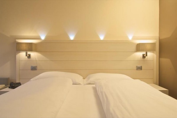 Dvoulůžková postel s dřevěným čelem a dvěma nástěnnými lampami.