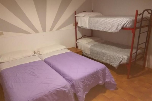 Dvě jednolůžka s fialovými přikrývkami a patrová postel v rohu pokoje s oranžovou podlahou.