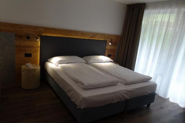 Moderní ložnice s manželskou postelí, dřevěnou podlahou a velkým oknem s průsvitnými závěsy.