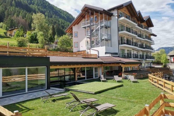 Moderní hotel s terasou a lehátky na svěží zelené zahradě obklopený horami.