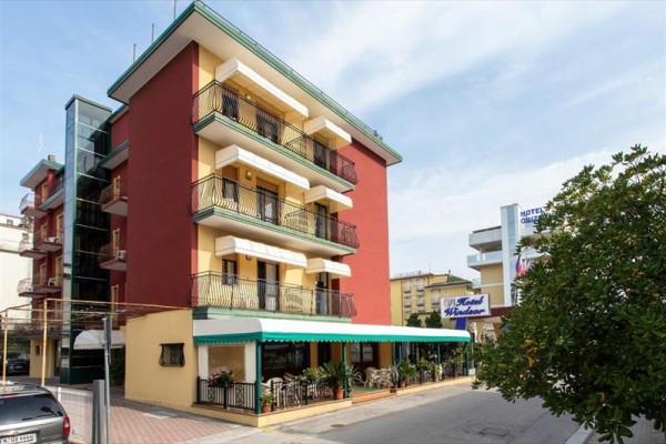 Čtyřpatrový hotel s verandou a balkonem, obklopený zelení a sousedními budovami.