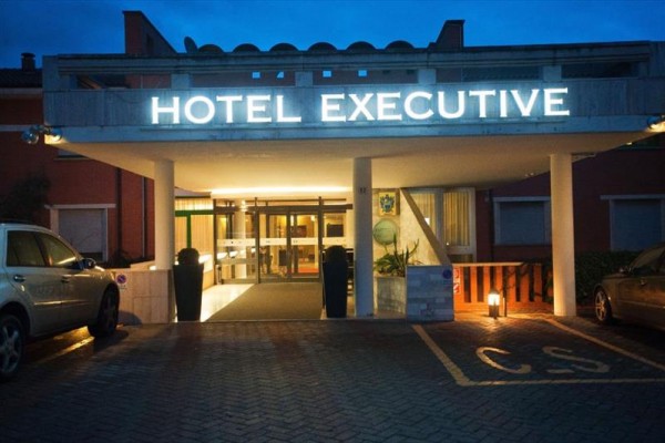Vchod do hotelu Executive s osvětleným nápisem a zaparkovanými auty.