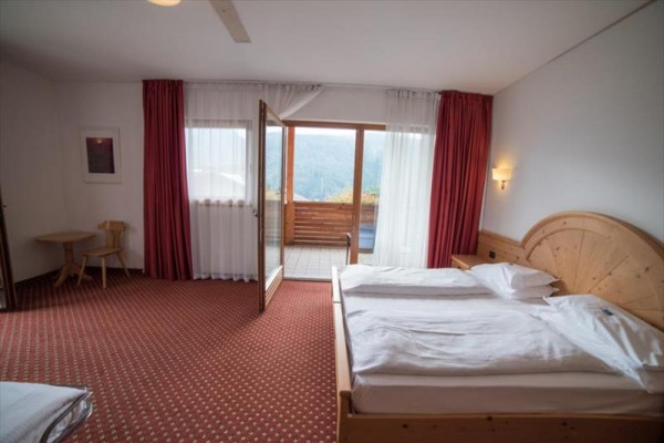 Hotelový pokoj s manželskou postelí, otevřenými dveřmi na balkon a výhledem na hory.