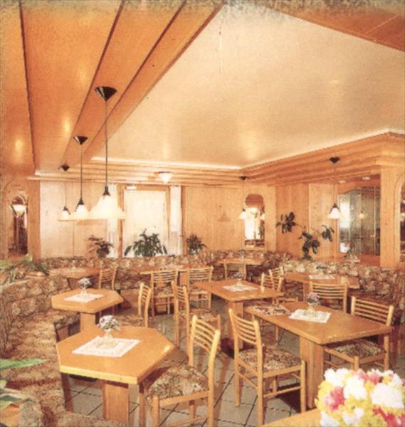 Restaurace s dřevěným nábytkem, rostlinami a lustry, se stoly a židlemi uspořádanými do řady.