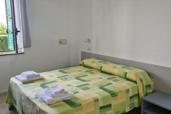Ložnice s otevřeným oknem, manželskou postelí se zelenožlutým přehozem a složenými ručníky.