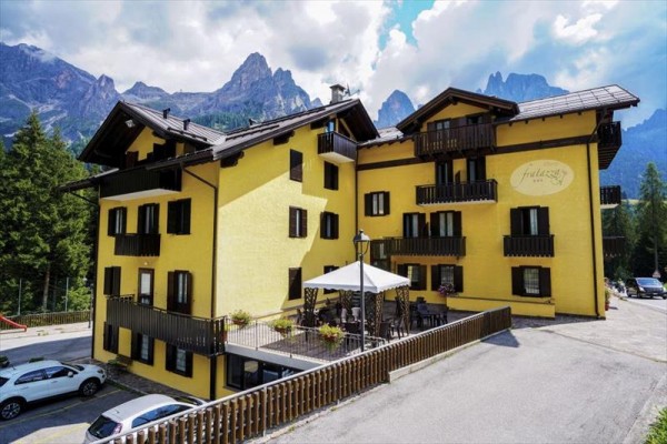 Žlutý hotel s dřevěnými balkony obklopený horami a lesy.