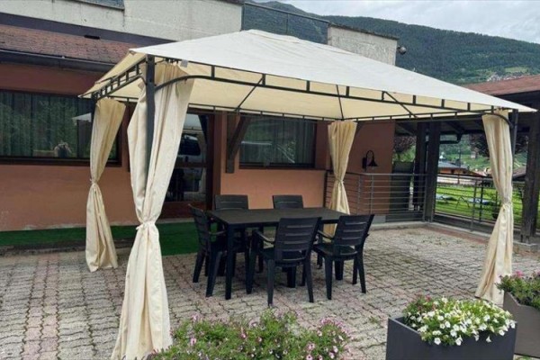 Stůl se šesti židlemi pod pergolou s bílou plachtou na terase, obklopený květinami.