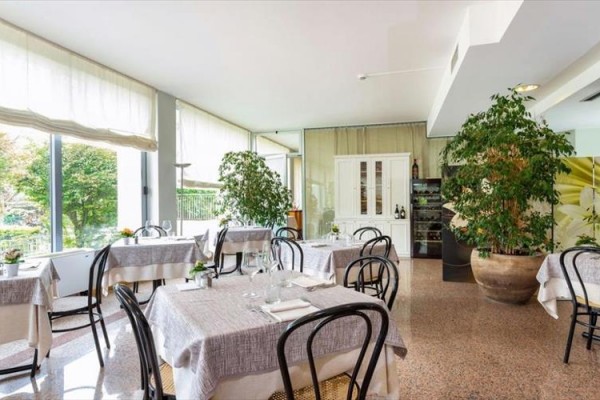 Restaurace s prostřenými stoly u velkých oken a zelenými rostlinami v květináčích.