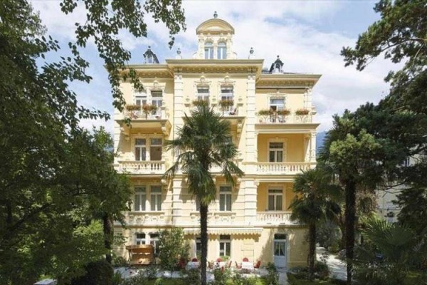 Historická vila obklopená zelení s palmovými stromy v popředí. 