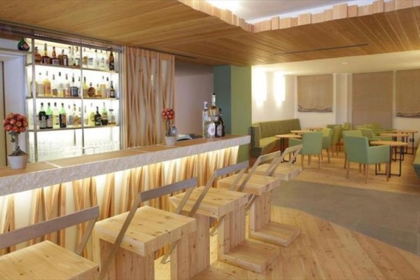 Moderní bar s dřevěnými židlemi a lavicemi, osvětlený pult s lahvemi alkoholu na policích v pozadí.