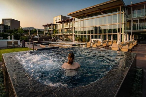 Osoba v lázních relaxuje ve vířivce před moderní budovou při západu slunce.