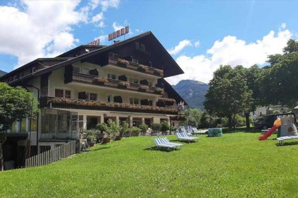 Hotel s balkonovou výzdobou a lehátky na trávníku, v pozadí hory.