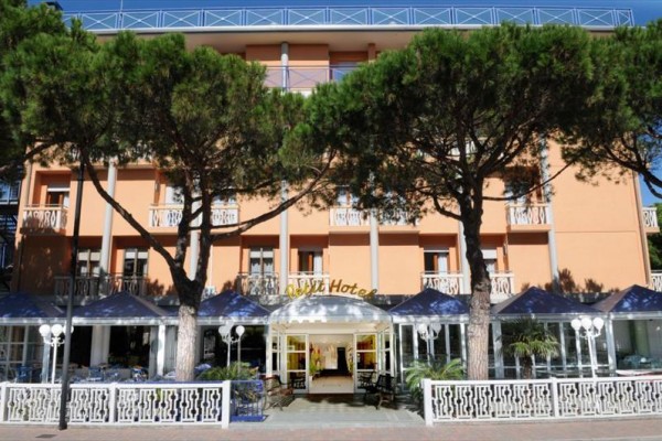 Vstup do hotelu s oranžovou fasádou, lemovaný stromy a modrými markýzami.