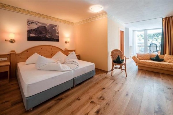 Prosvětlený hotelový pokoj s manželskou postelí, rozkládací pohovkou a dřevěnými podlahami.