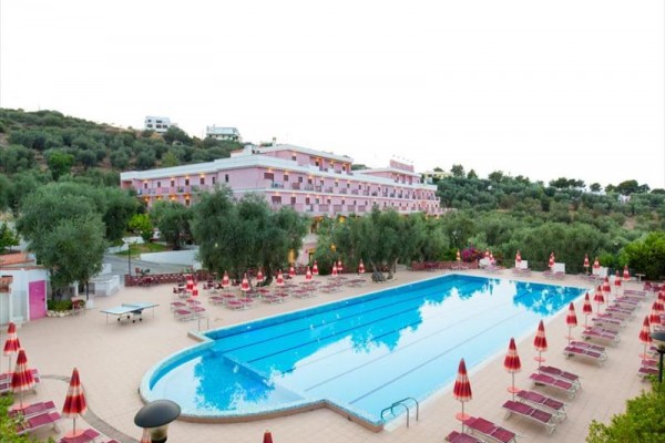 Bazén s lehátky a slunečníky před růžovou budovou hotelu obklopenou zelení.
