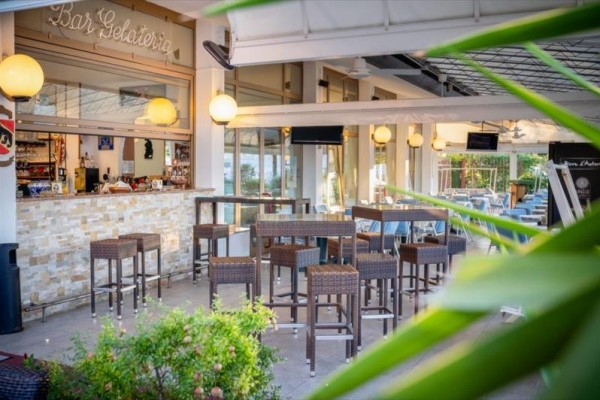 Venkovní bar s vysokými stolky a židlemi, ozdobený lampami a rostlinami.