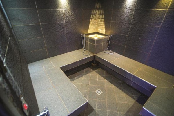 Parní sauna s lavicemi a sprchovými hlavicemi na tmavé dlažbě.