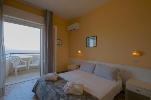 Pokoj s manželskou postelí, nočními stolky a výhledem na balkon s plastovými židlemi.
