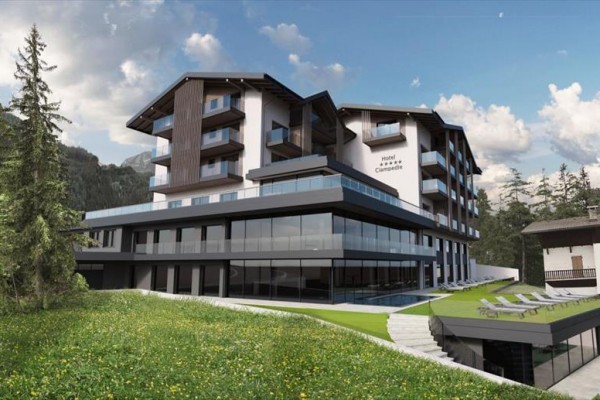 Moderní hotel obklopený horami a lesy s prosklenými terasami a relaxačními lehátky na zeleném trávníku.